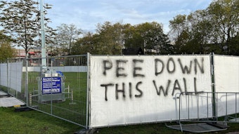 Auf einem Transparent, welches an einem Bauzaun vor den Uniwiesen hängt, steht „Pee Down This Wall“