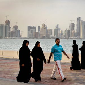 Zwei katarische Frauen und ein Mann spazieren vor der Skyline von Doha. Im Hintergrund spazieren drei Frauen. Alle Frauen tragen lange, schwarze Kleider und Kopftücher. Der Mann trägt eine weiße Hose und ein hellblaues Hemd.