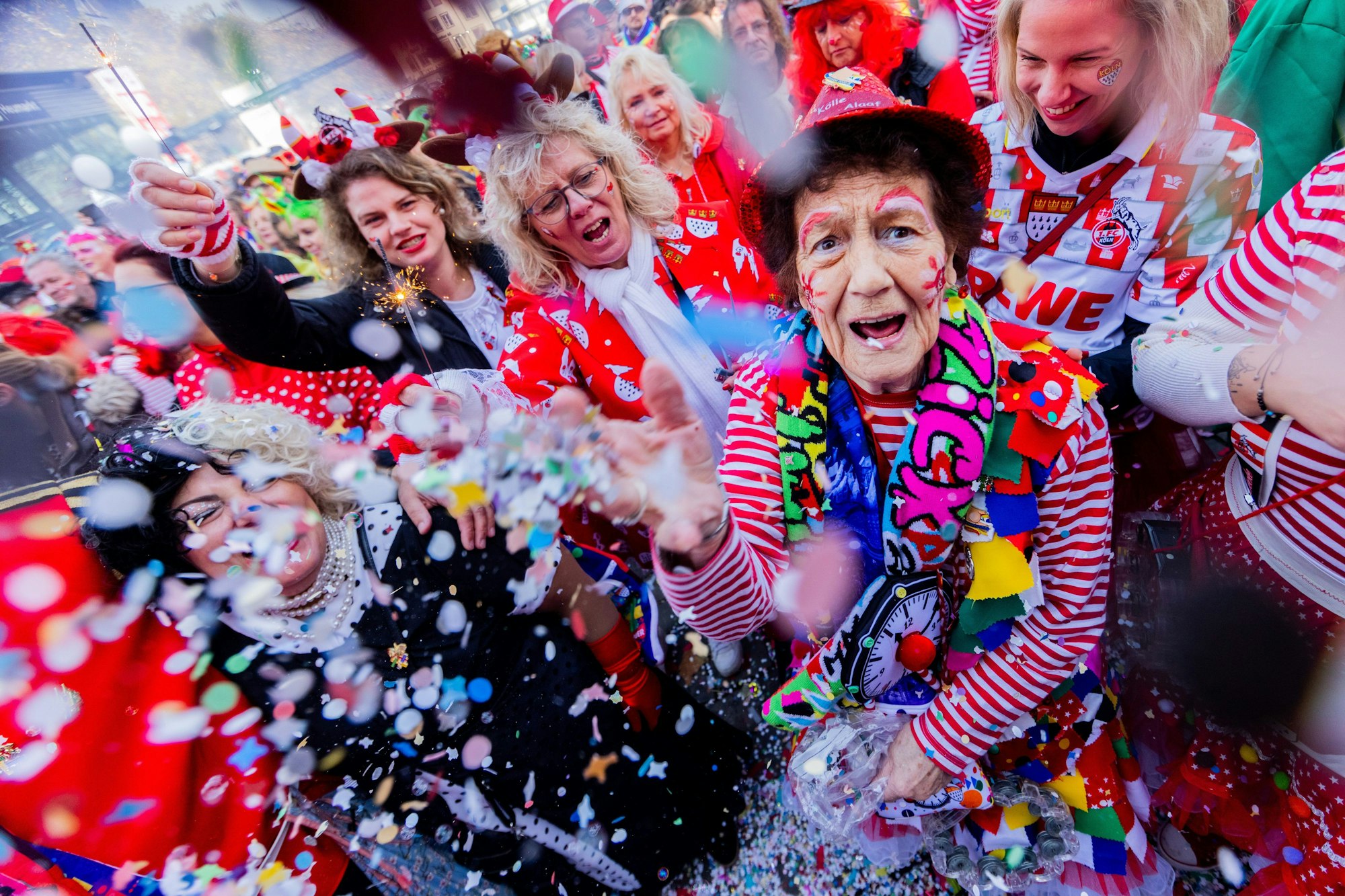Jecken feiern den Auftakt der Karnevalssession auf dem Heumarkt. Eine ältere Frau blickt in die Kamera und wirft Konfetti.