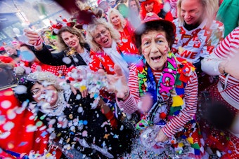 Jecken feiern den Auftakt der Karnevalssession auf dem Heumarkt. Eine ältere Frau blickt in die Kamera und wirft Konfetti.