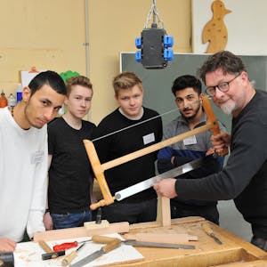 Ausbilder Arnulf Rückamp steht mit seinen Lehrlingen um einen Tisch mit Werkzeug und sägt Holz.