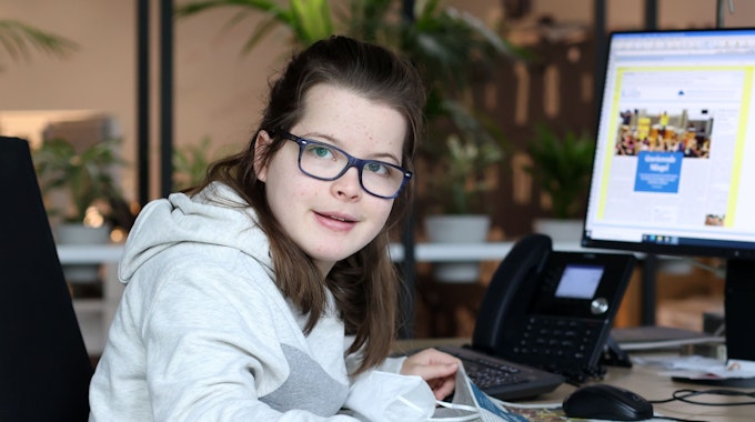 Die gehandicapte Schülerin Aimée Heimbach sitzt an einem Schreibtisch in der Lokalredaktion des Kölner Stadt-Anzeigers vor einem großen Monitor, auf dem die aktuelle Zeitungsseite zu sehen ist.&nbsp;