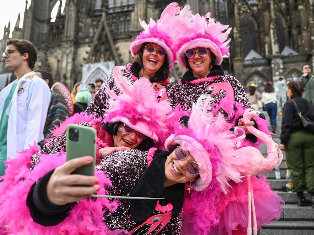 Verkleidete Besucher machen ein Selfie vor dem Kölner Dom. Sie haben ein pinkes Kostüm mit zahlreichen Federn.