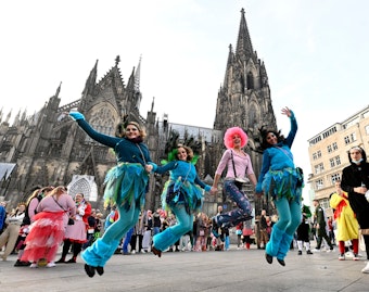 Eine Gruppe Karnevalisten springt vor dem Dom in die Höhe. Sie haben teilweise pastellblaue Hosen, Röcke und Oberteile an.