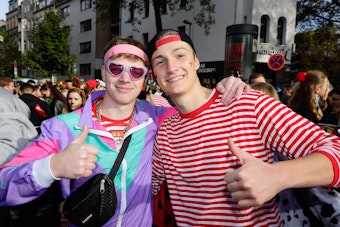 Zwei junge Männer auf der Zülpicher Straße. Einer hat einen bunten Trainingsanzug an, der rechte ist mit einem rot-weißen Streifenshirt bekleidet.