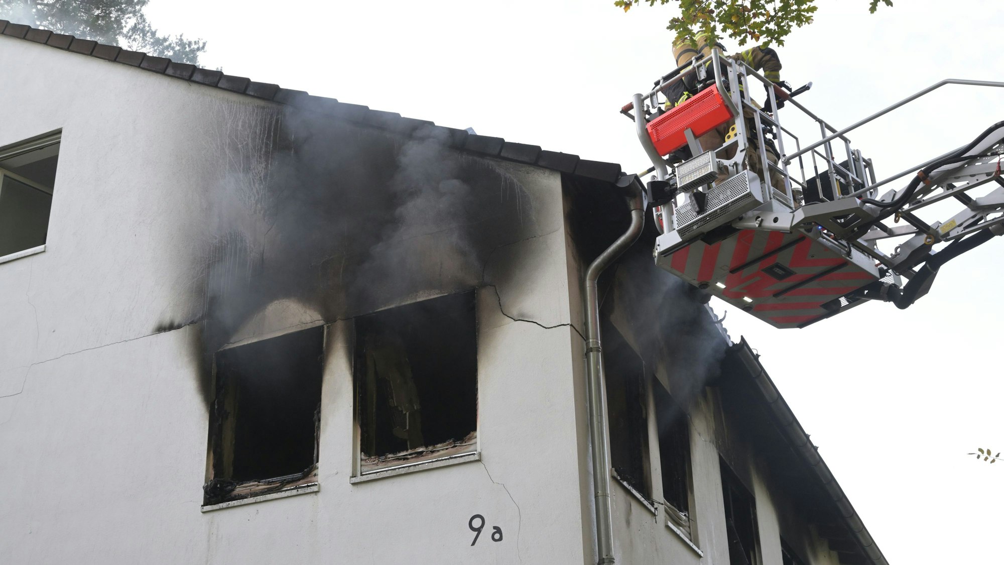 Ein Feuerwehrmann steht in einem Korb auf der Feuerwehrleiter. Die Hausfassade ist an mehreren Fenstern von dem Feuer zerstört.