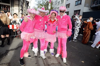 Vier Männer in pinken Kostümen und ebenfalls rosafarbenen Hüten stehen auf der Straße nebeneinander.