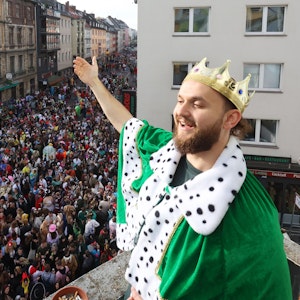 Ein junger Mann steht mit einer Krone auf dem Balkon. Unter ihm die volle Zülpicher Straße.