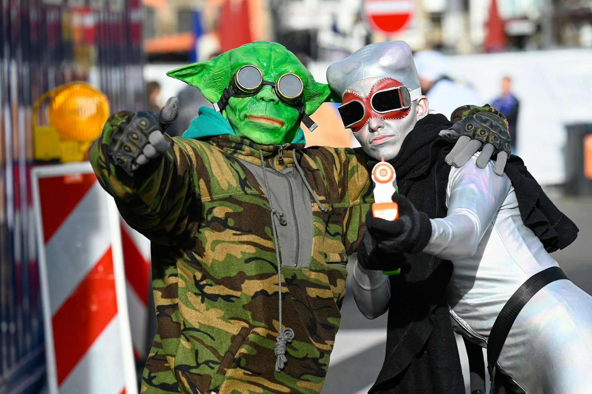 Zwei Männer sind als Außerirdische verkleidet. Einer trägt eine grüne Alien-Maske, der andere hat eine silberne Kopfbedeckung auf und trägt eine Waffe in der Hand.