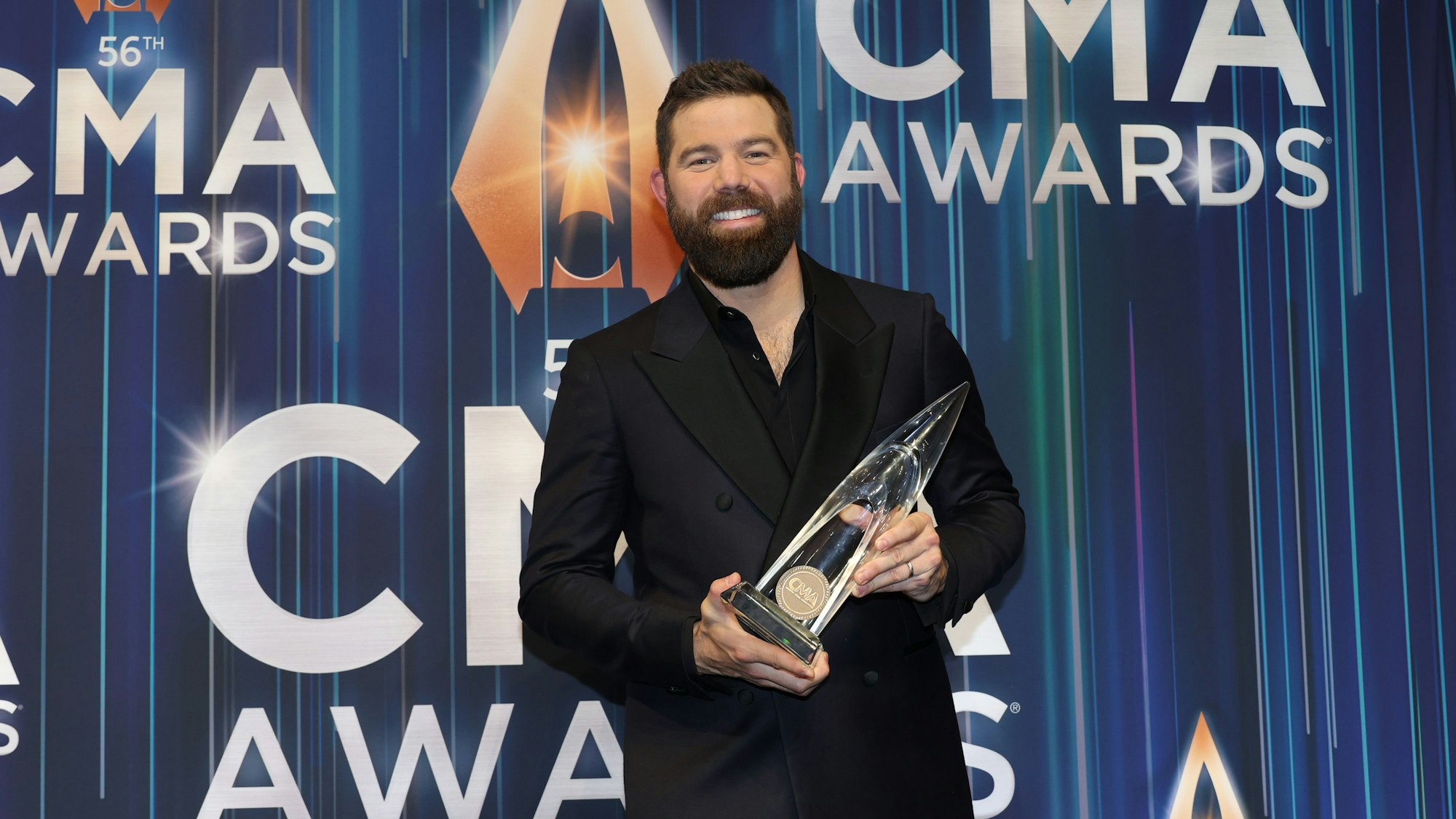 Jordan Davis mit seinem CMA Award für den Song des Jahres. Er lacht. Er trägt einen schwarzen Anzug und Vollbart.