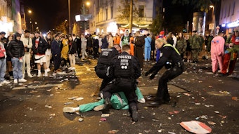 Auf der Zülpicher Straße liegt ein Karnevalist auf der Straße, drei Polizei-Beamte nehmen in fest. Drumherum stehen viele Menschen.



