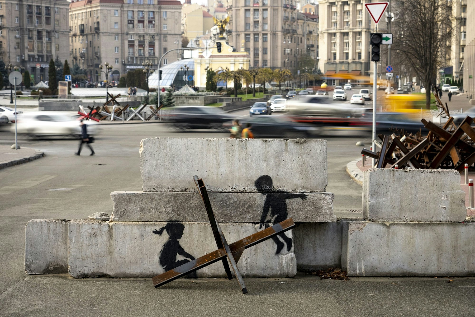 Streetart: Zwei gemalte Kinder sind auf einem Betonblock zu sehen, im Vordergrund ist ein Metallhindernis. Es sieht so aus, als würden die Kinder auf dem Hindernis schaukeln.