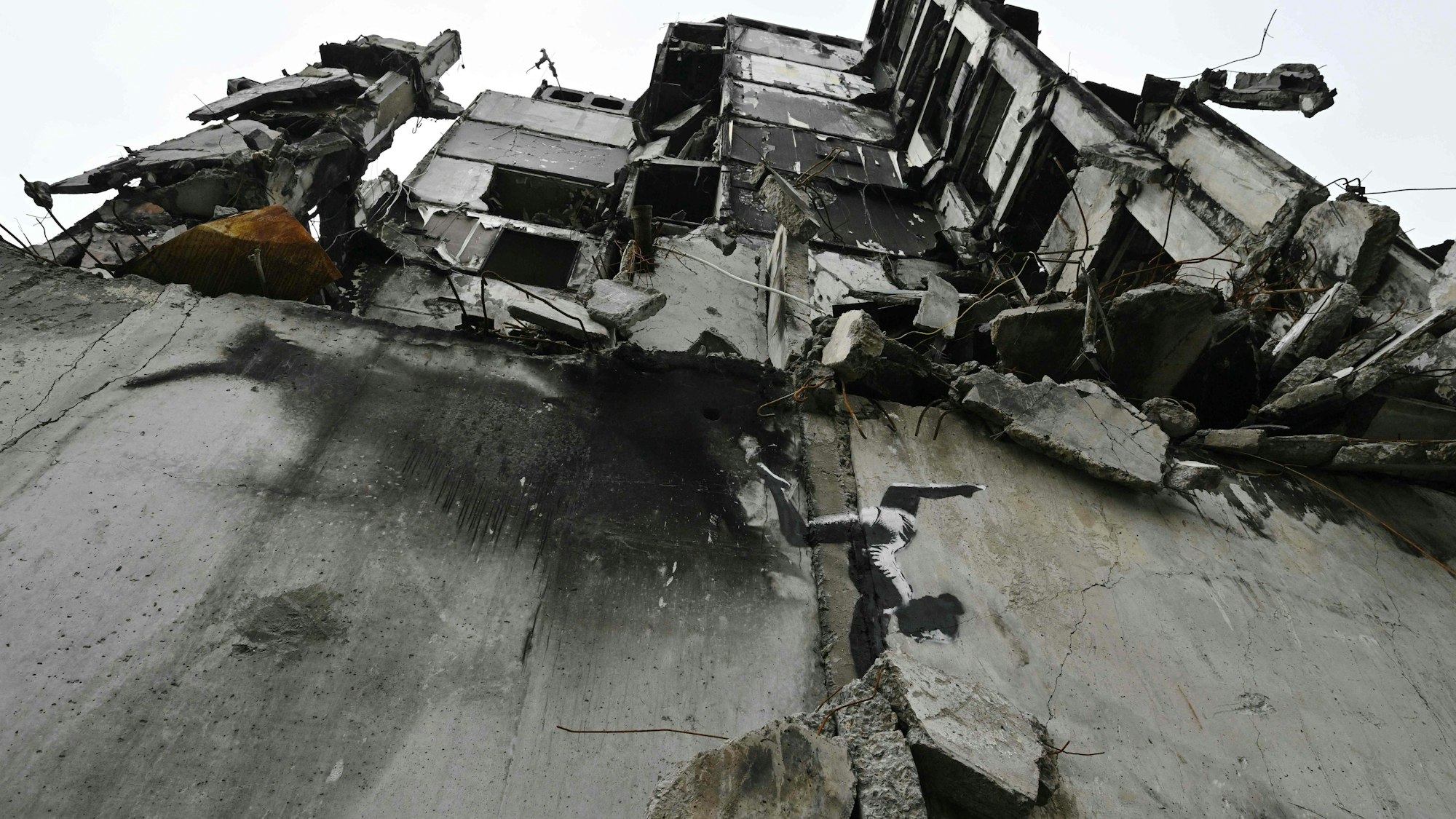 Ein vom Krieg zerstörtes Haus in der Ukraine ist zu sehen, im Vordergrund ist auf einer Betonmauer eine gemalte Tänzerin zu sehen, die einen Handstand macht.