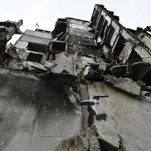 Ein vom Krieg zerstörtes Haus in der Ukraine ist zu sehen, im Vordergrund ist auf einer Betonmauer eine gemalte Tänzerin zu sehen, die einen Handstand macht.