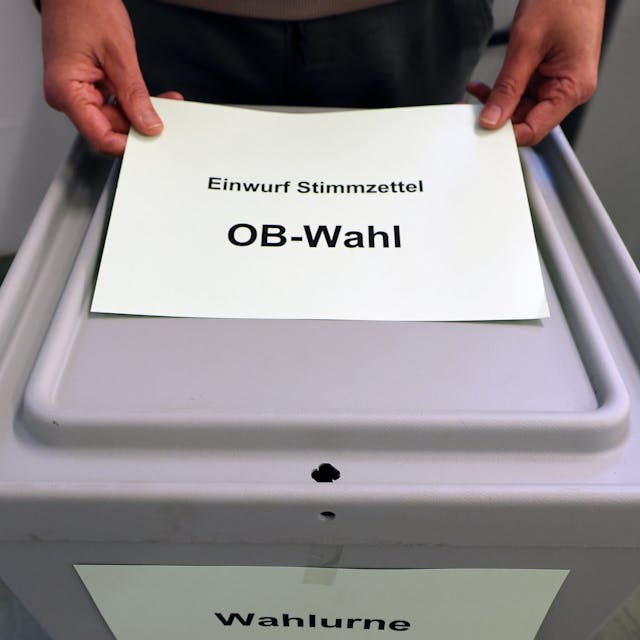 Ein Zettel mit der Aufschrift „Einfwurf Stimmzettel OB-Wahl“ wird vor einer grauen Wahlurne gezeigt.