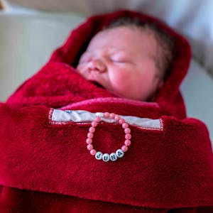 Ein neugeborenes Kind liegt in eine rote Decke eingewickelt in einem Bett. Auf der Decke liegt eine Kette mit dem Namen des Kindes: Juna.