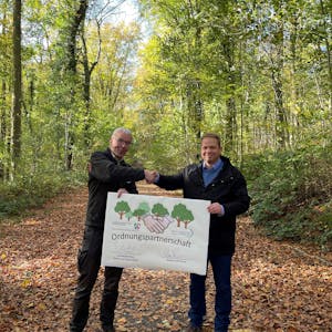 Forstamtsleiter Stephan Schütte und Uwe Zaar, Kreisdezernent Umwelt, Planung und Verkehr, halten im Königsdorfer Forst den Vertrag hoch, mit dem die Zusammenarbeit besiegelt wurde.