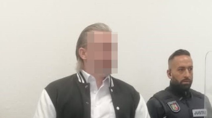 Der Angeklagte steht hinter einem Stuhl im Gerichtssaal. Er trägt eine schwarz-weiße Jacke, neben ihm steht ein Justiz-Beamter. Das Gesicht des Angeklagten ist gepixelt.
