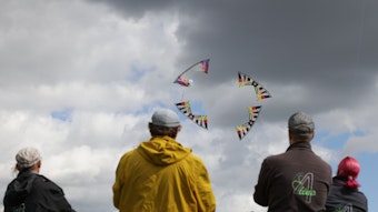 vier Personen stehen mit dem Rücken zum Betrachter und schauen auf Drachen, die vor einem wolkigen Himmel fliegen