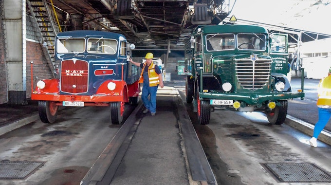 Zwei LKW aus den 1950er-Jahren stehen in der Brikettfabrik in Frechen und warten, dass sie mit Kohle beladen werden. Ein Arbeiter steht mit Helm und Warnweste neben dem linken LKW.