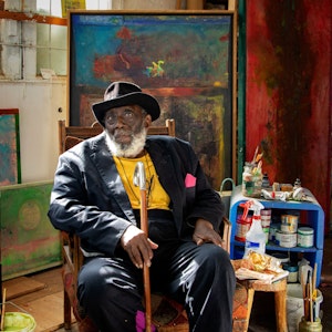 Der Künstler Frank Bowling sitzt in seinem Atelier vor Leinwänden und Malutensilien.