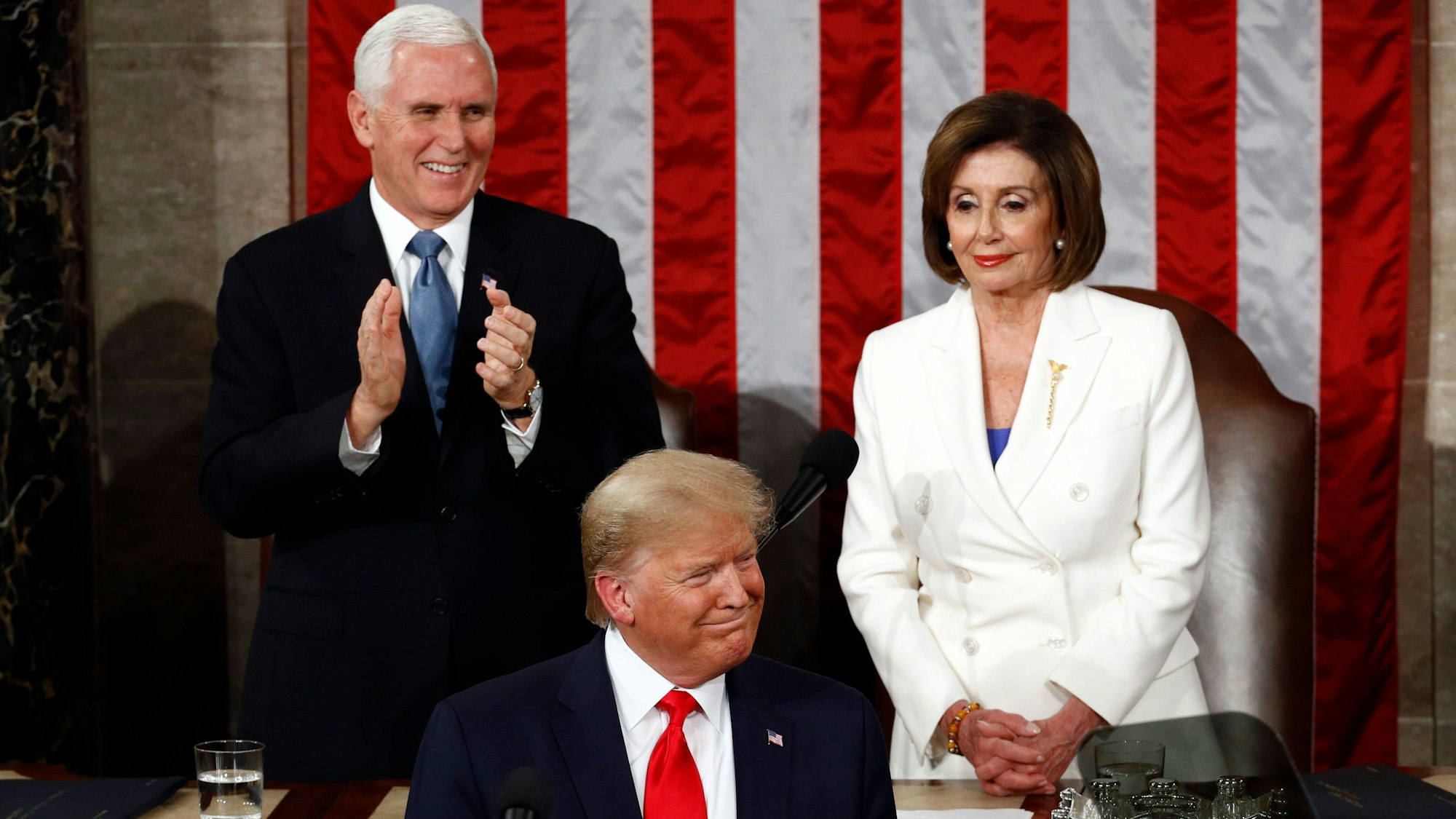 Donald Trump steht im Vordergrund vor einem Mikrofon, im Hintergrund applaudiert Mike Pence, Nancy Pelosi hat die Hände gefaltet. An der Wand hängt eine große Flagge der USA.