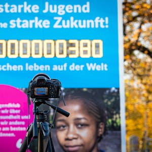 Es ist eine Kamera zu sehen, die auf die Weltbevölkerungsuhr zeigt. Die Zahl auf dieser hat acht Milliarden erreicht.