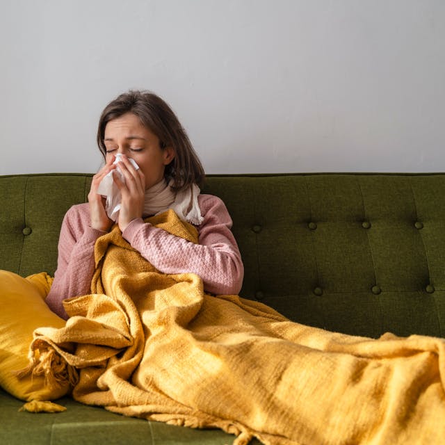Eine Frau liegt mit einer Decke und einem Schal auf einem Sofa und putzt sich mit einem Taschentuch die Nase.