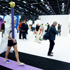Ein Performance-Künstler mit weißer Schleppe und Pfosten geht durch eine Messehalle der Kunstmesse Art Cologne.&nbsp;