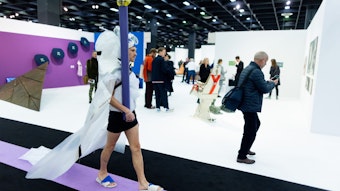 Ein Performance-Künstler mit weißer Schleppe und Pfosten geht durch eine Messehalle der Kunstmesse Art Cologne.