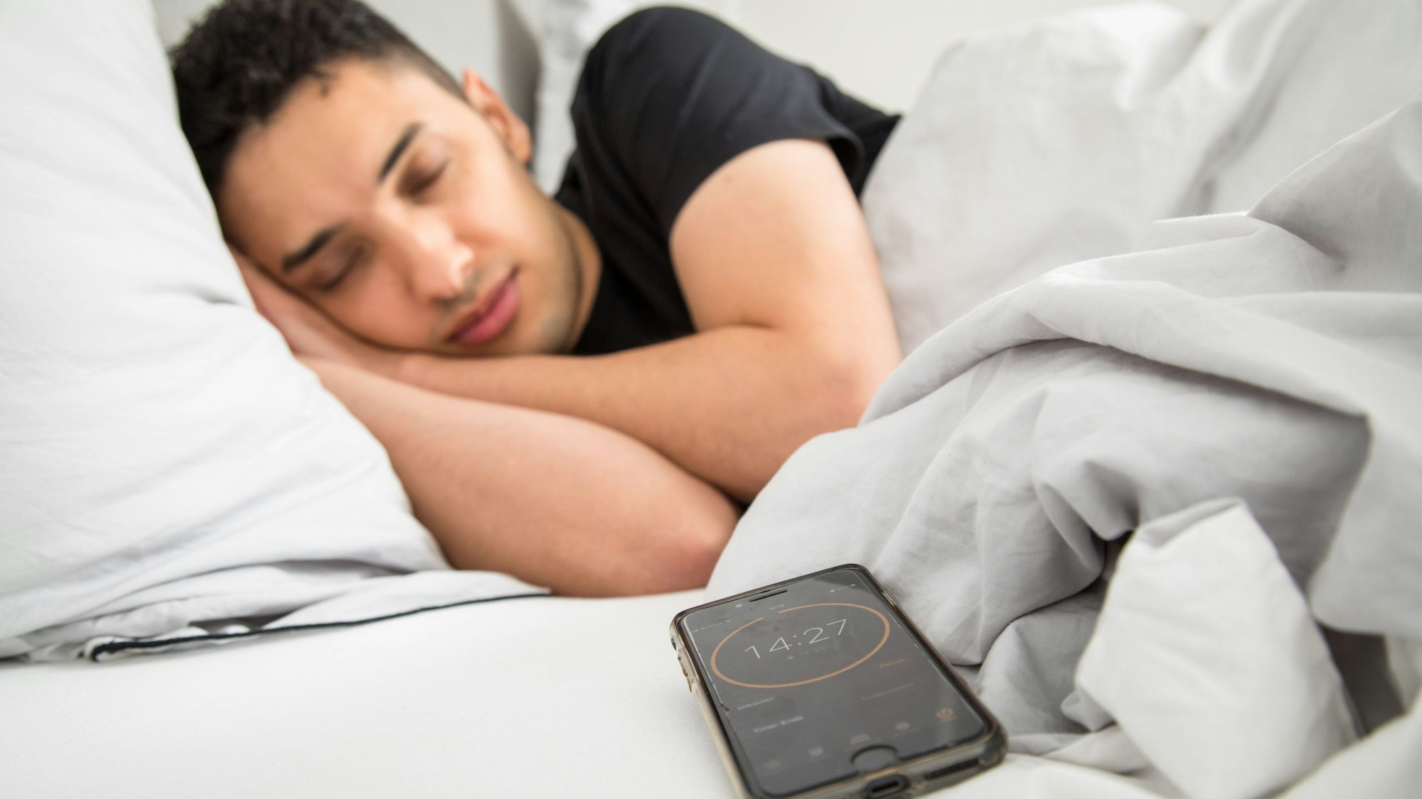 Ein junger Mann schläft im Bett, im Vordergrund ist sein Smartphone mit gestelltem Timer zu sehen.