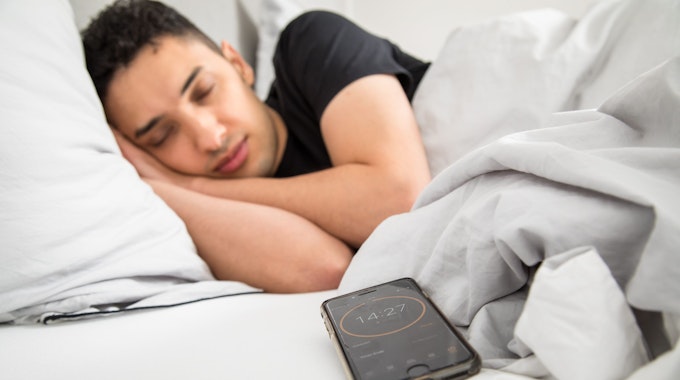 Ein junger Mann schläft im Bett, im Vordergrund ist sein Smartphone mit gestelltem Timer zu sehen.