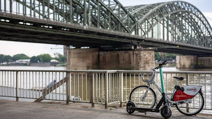 Ein E-Scooter und ein Leihfahrrad der Kölner Verkehrsbetriebe (KVB) stehen am Rheinufer vor der Hohenzollernbrücke in der Kölner Altstadt.