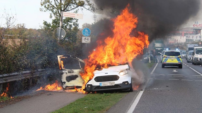 Hohe Flammen schlagen aus einem Auto, das direkt an einer Straße steht.