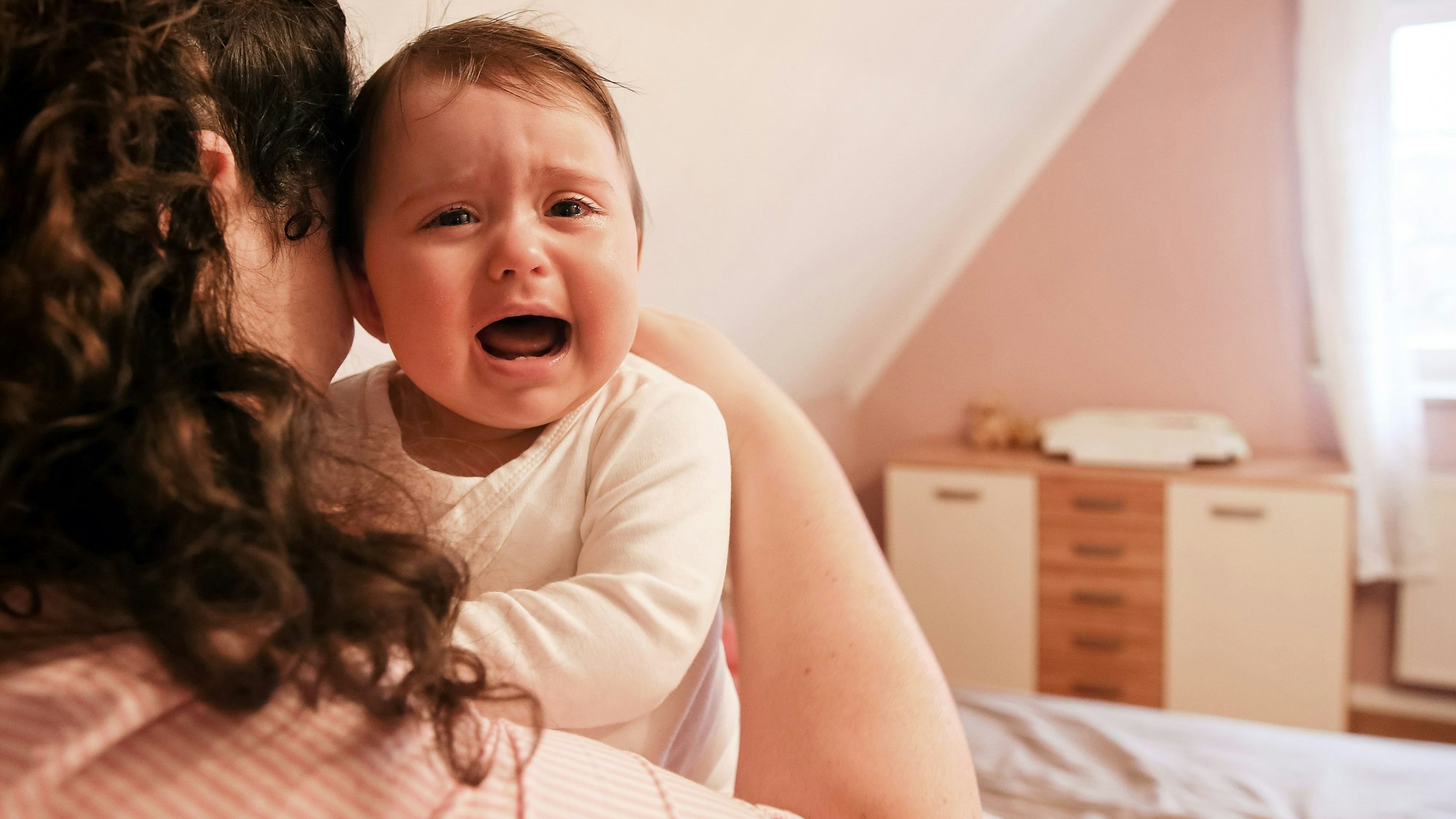 Eine Mutter ist von hinten im Bett sitzend zu sehen, sie hält ein weinendes Baby auf dem Arm, das in die Kamera guckt.
