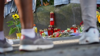 Menschen stehen vor einem Zaun, an dem mit Kerzen und Blumen eines von der Polizei erschossenen Jugendlichen gedacht wird.