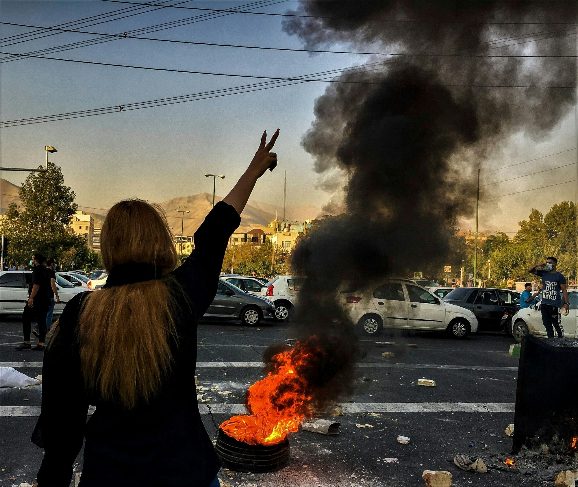 30.09.2022, Iran, Teheran: Eine Frau steht während einer Demonstration nach dem Tod der 22-jährigen Mahsa Amini vor einem brennenden Autoreifen und zeigt das Victory-Zeichen.