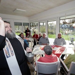 Pater Georg segnet einen Raum mit Weihwasser. Im Hintergrund sitzen Seniorinnen und Senioren an Tischen vor einer Fensterfront.
