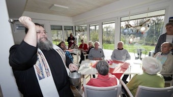 Pater Georg segnet einen Raum mit Weihwasser. Im Hintergrund sitzen Seniorinnen und Senioren an Tischen vor einer Fensterfront.