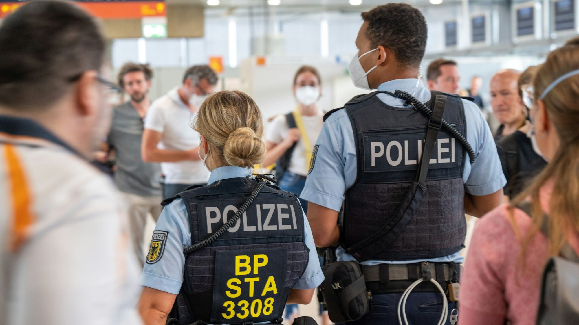 Zu sehen sind zwei Polizisten in der Rückansicht am Flughafen Köln/Bonn. Daneben, davor und dahinter befinden sich Fluggäste.