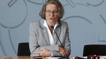 NRW-Ministerin Ina Scharrenbach (CDU) bei einer Pressekonferenz. Sie sitzt an einem Tisch, vor ihr ein schmales Mikrofon und hinter ihr eine graue Wand.