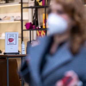 Ein Schild am Eingang eines Schuh-Geschäfts weist auf die Pflicht zum Tragen einer FFP2-Maske hin, während im Vordergrund eine Frau mit FFP2-Maske vorbeigeht.
