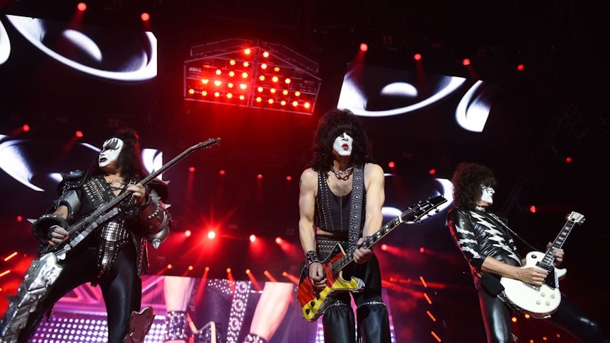 Drei Mitglieder der Band Kiss mit Gitarren auf der Bühne bei einem Konzert.