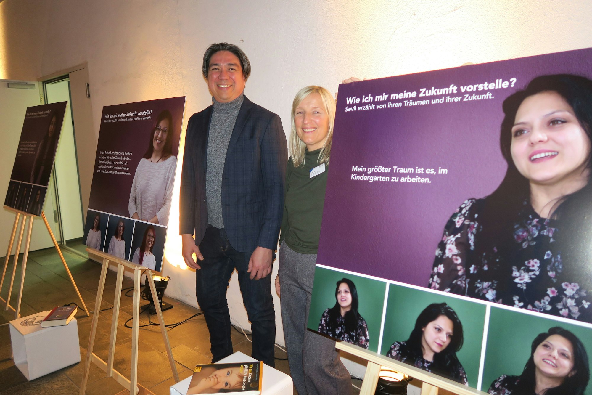 Jhon Jimenez und Anna Drahs in der Ausstellung. Neben ihm sind Plakate aufgestellt, auf denen Bilder von jungen Frauen zu sehen sind. Neben den Frauen sind Zitate von diesen dargestellt.