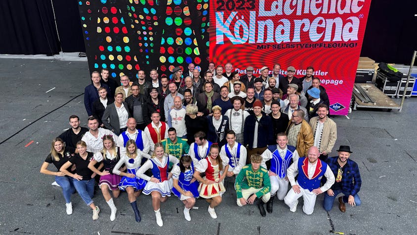 Rund 50 Mitwirkende der Lachenden Kölnarena 2023, Topbands des Kölner Karnevals und Tanzgruppenmitglieder, präsentieren sich vor einem digitalen Großplakat.&nbsp;&nbsp;