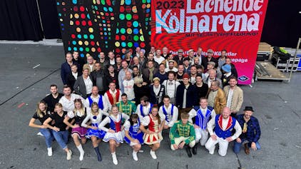 Rund 50 Mitwirkende der Lachenden Kölnarena 2023, Topbands des Kölner Karnevals und Tanzgruppenmitglieder, präsentieren sich vor einem digitalen Großplakat.&nbsp;&nbsp;
