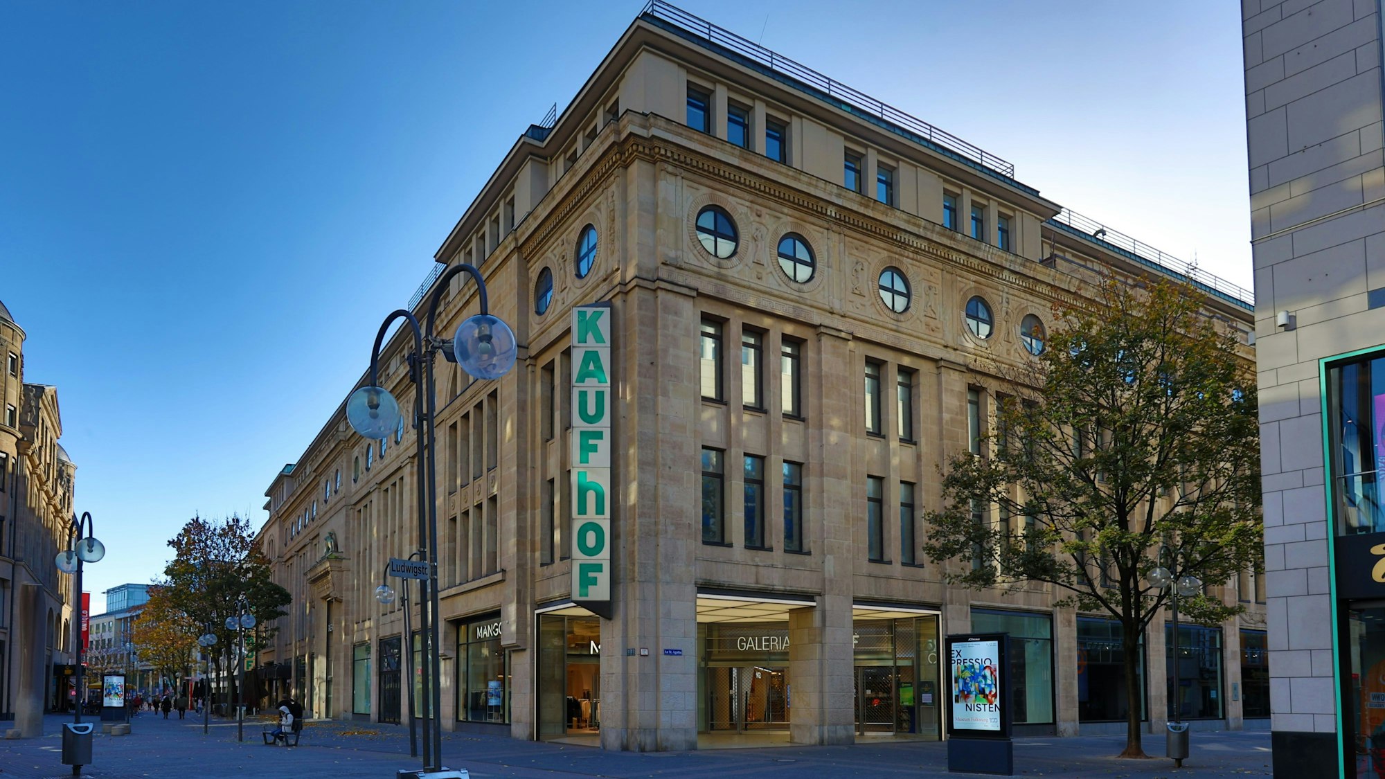 Außenansicht des viergeschossigen Galeria Karstadt Kaufhof-Gebäudes in der Kölner Schildergasse. Die helle Sandsteinfassade hebt sich vom blauem Himmel ab, der über zwei Etagen reichende Schriftzug „Kaufhof“ziert eine Gebäudeecke.