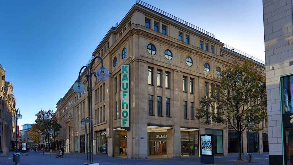 Außenansicht des viergeschossigen Galeria Karstadt Kaufhof-Gebäudes in der Kölner Schildergasse. Die helle Sandsteinfassade hebt sich vom blauem Himmel ab, der über zwei Etagen reichende Schriftzug „Kaufhof“ ziert eine Gebäudeecke.