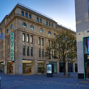 Außenansicht des viergeschossigen Galeria Karstadt Kaufhof-Gebäudes in der Kölner Schildergasse. Die helle Sandsteinfassade hebt sich vom blauem Himmel ab, der über zwei Etagen reichende Schriftzug „Kaufhof“ ziert eine Gebäudeecke.