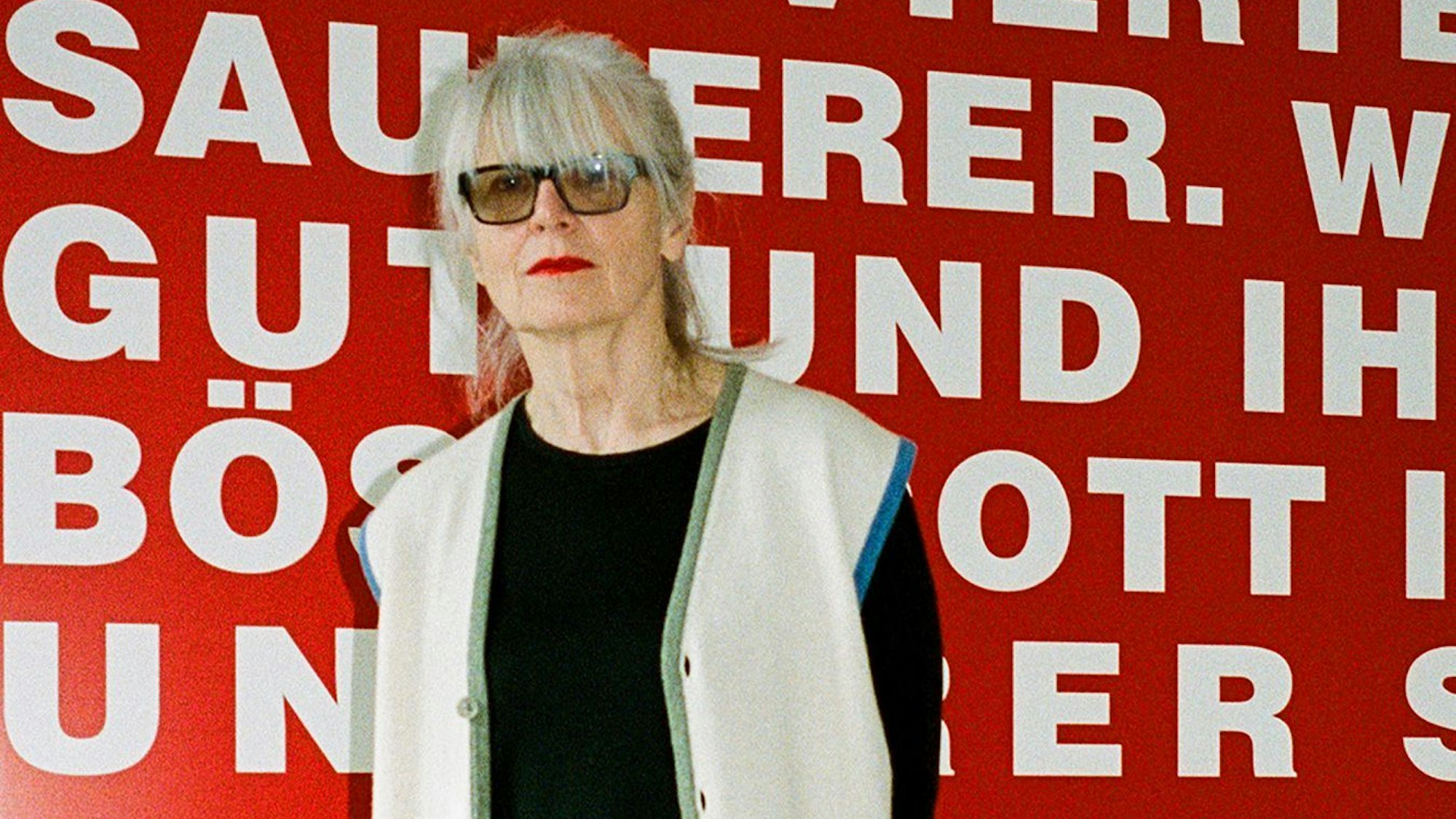 Die Galeristin Monika Sprüth steht vor einer roten Wand mit weißer Schrift.